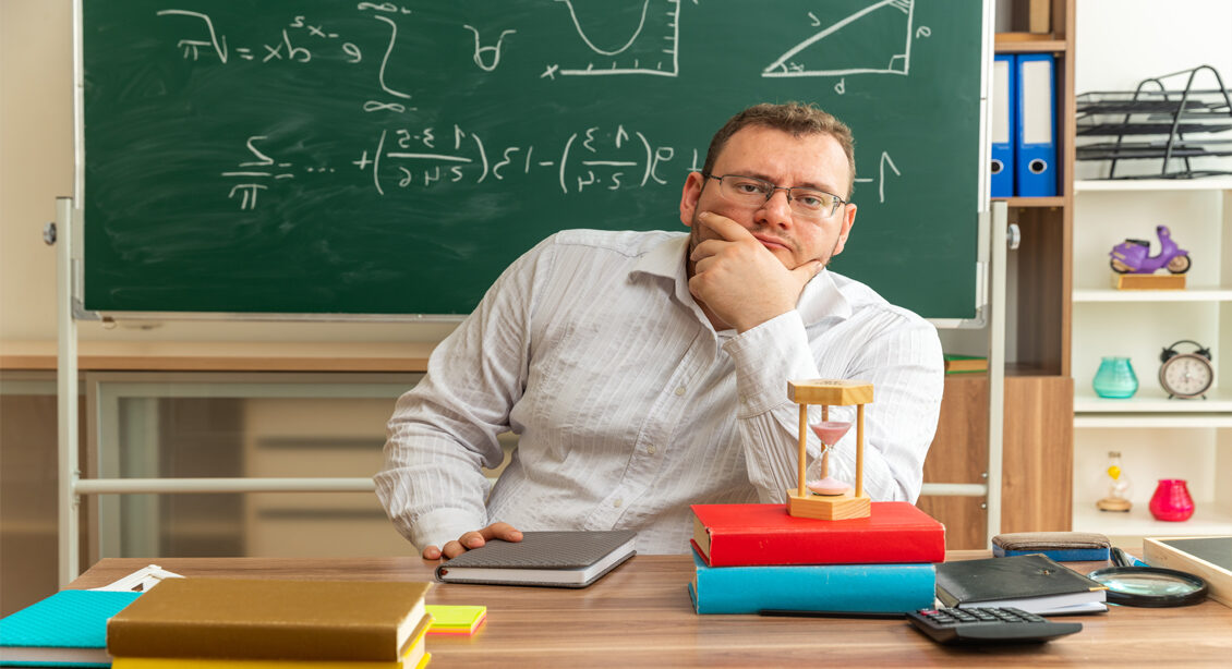 thoughtful-young-blonde-teacher-wearing-glasses-sitting-desk-with-school-tools-classroom-keeping-hand-chin-looking-camera